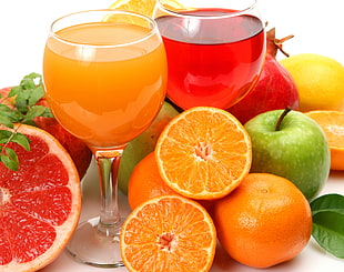 two footed clear drinking glasses filled with red and orange liquids surrounded with fruits